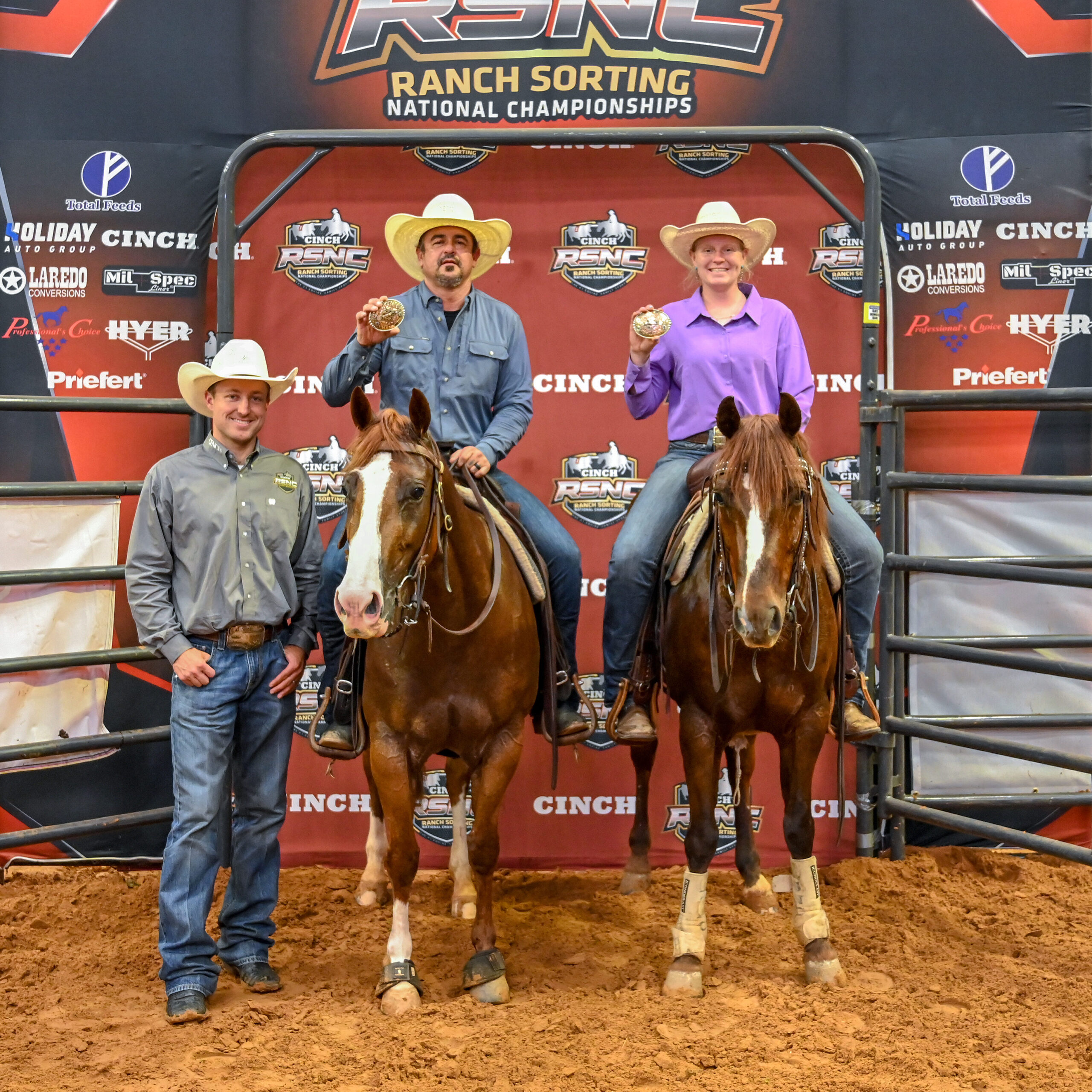 Two mounted riders hold up championship buckles beside a standing man in a cowboy hat at the Cinch RSNC backdrop