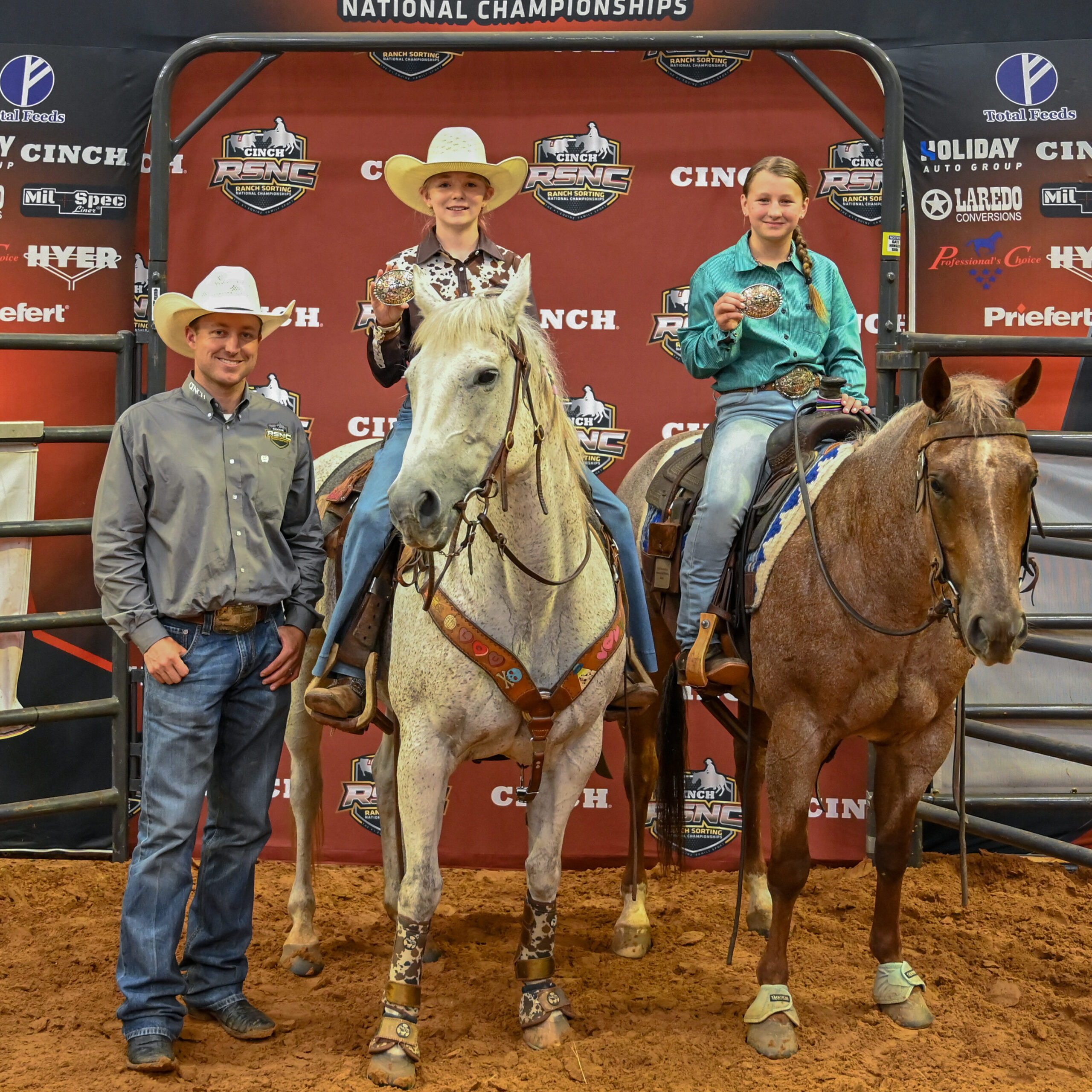 Two young riders hold up championship buckles on horseback beside a man in a cowboy hat at the Cinch RSNC backdrop