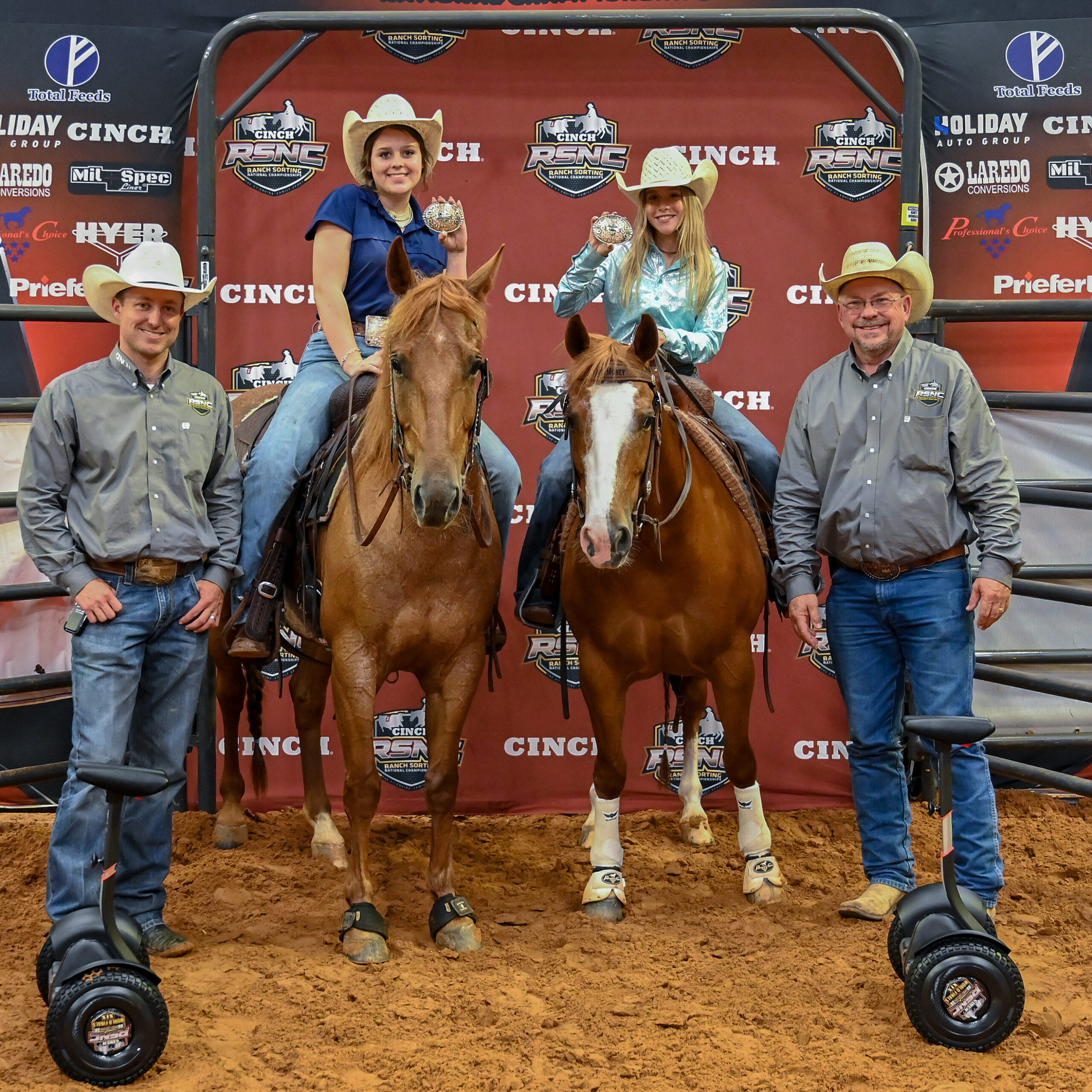 Two young women on horseback hold championship buckles between two men on Segways at a Cinch RSNC ranch sorting awards presentation