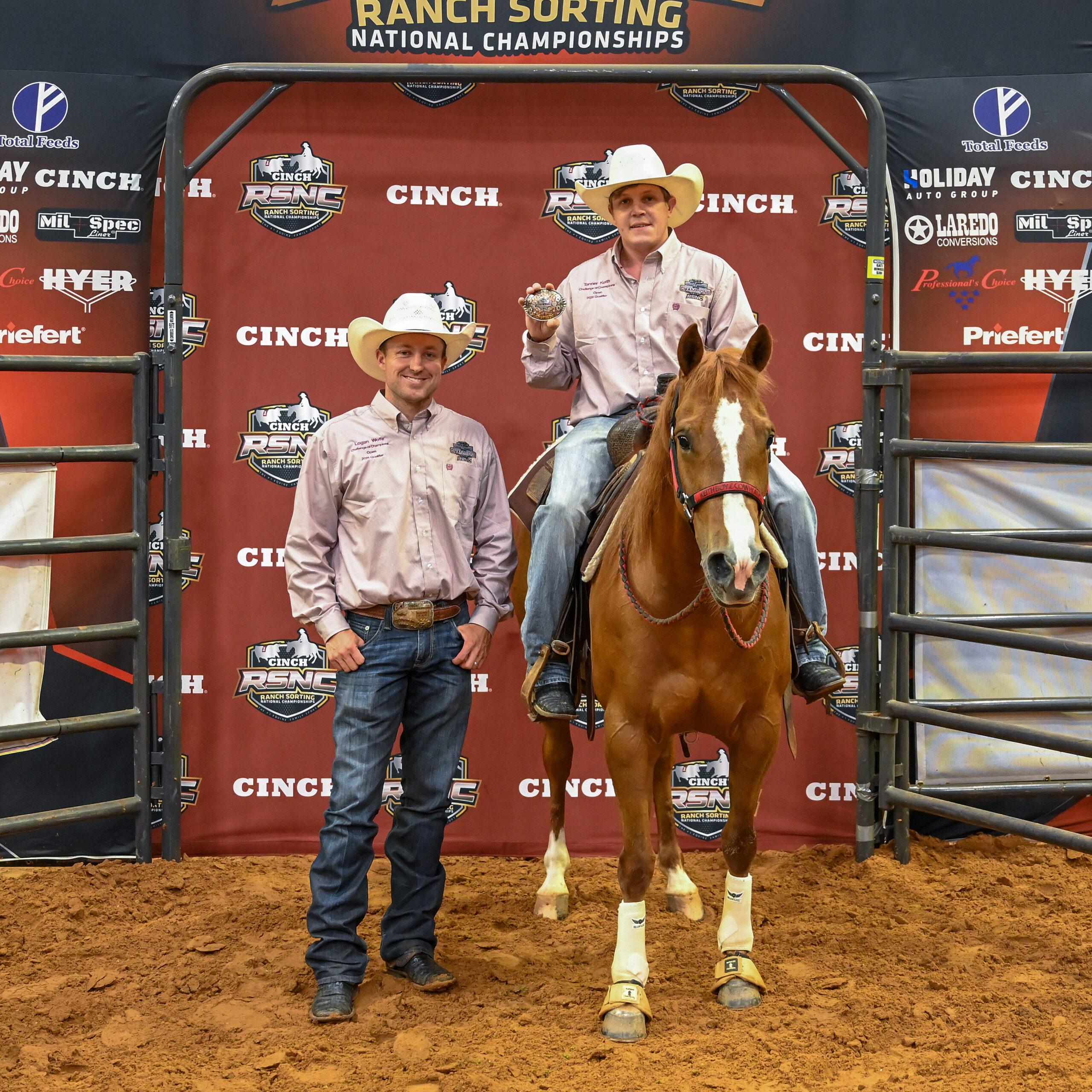 Two cowboys in matching shirts pose at the Cinch RSNC backdrop, one mounted on a sorrel horse holding up a championship buckle