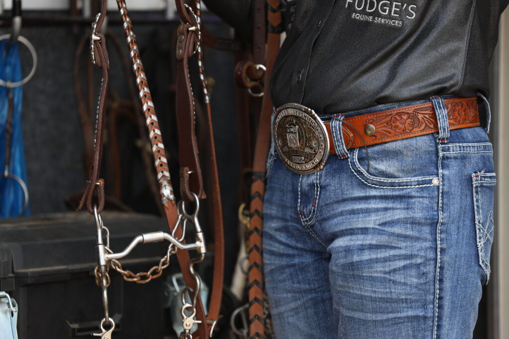 Close-up of a cowboy's tooled leather belt and large silver trophy buckle over jeans, with bridles and a bit hanging nearby