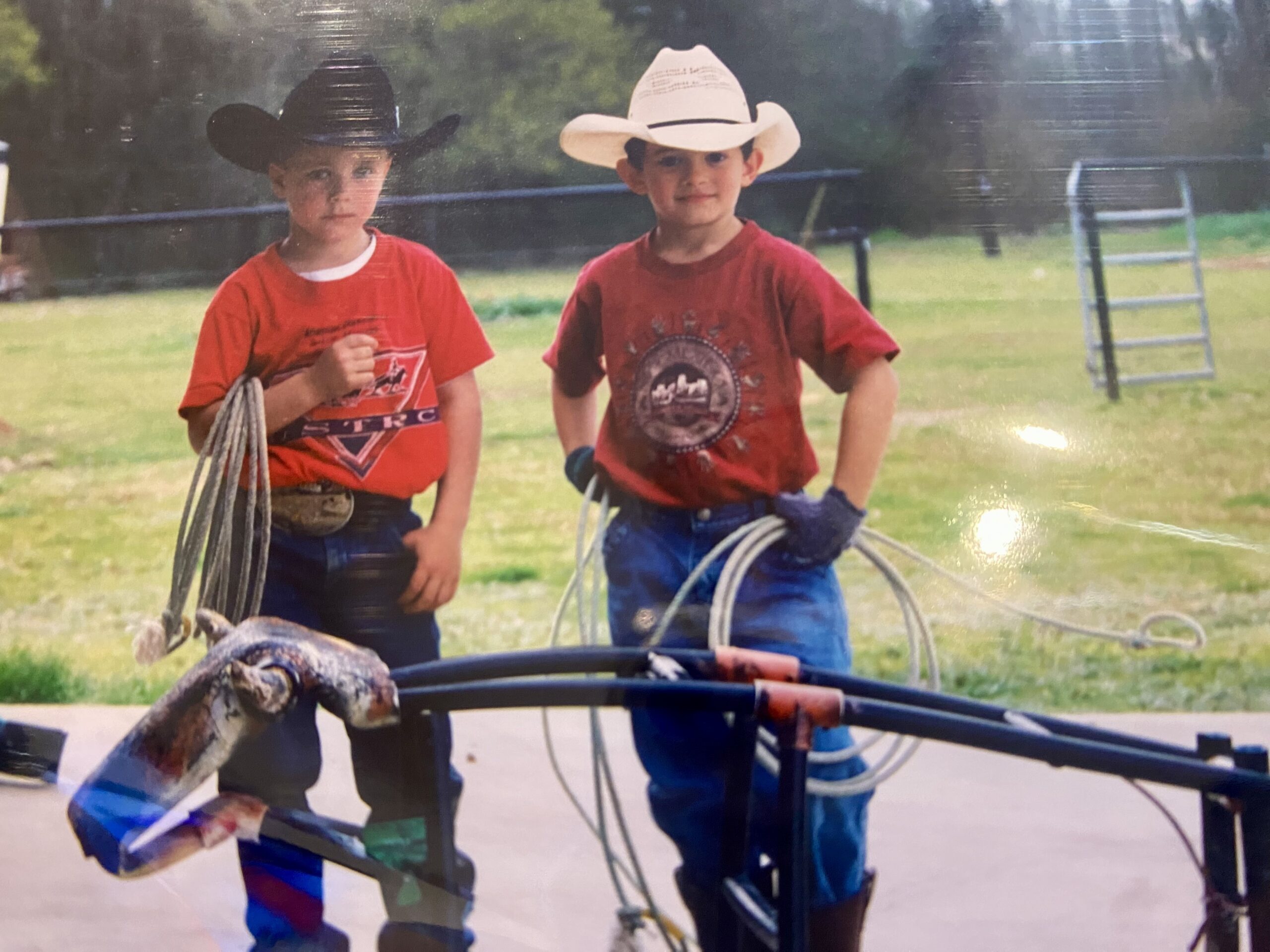 Two young boys in cowboy hats and red shirts hold coiled ropes beside a roping dummy on a grassy practice lot