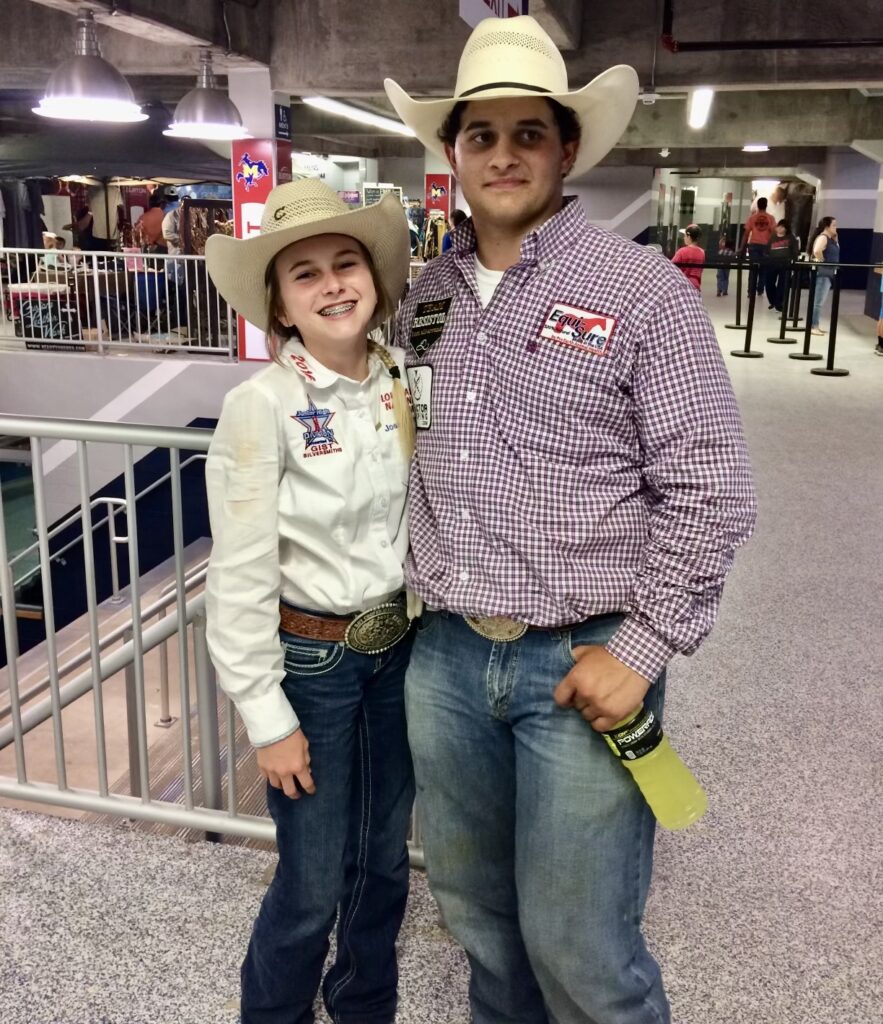 Two young rodeo contestants in cowboy hats, trophy buckles and contestant numbers pose side by side in an arena concourse