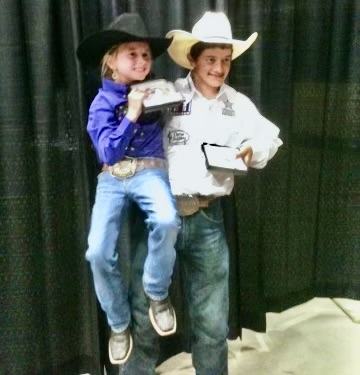 Two young rodeo competitors in cowboy hats and belt buckles stand side by side holding their prize buckle boxes
