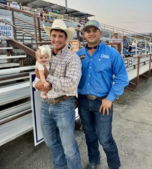 Two cowboys in trophy buckles stand by the rodeo arena bleachers, one holding a baby girl in a white bow