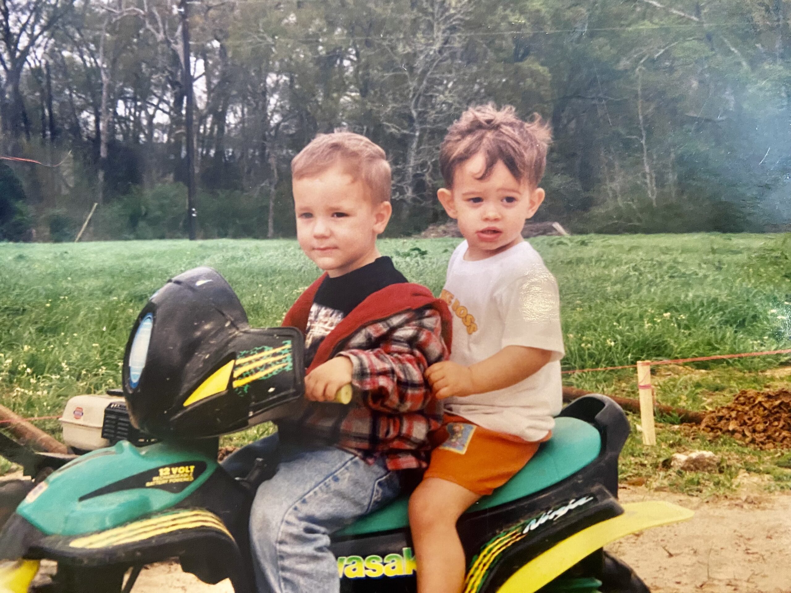 Two young boys sit together on a green battery-powered toy Kawasaki Ninja motorcycle in front of a grassy field and trees