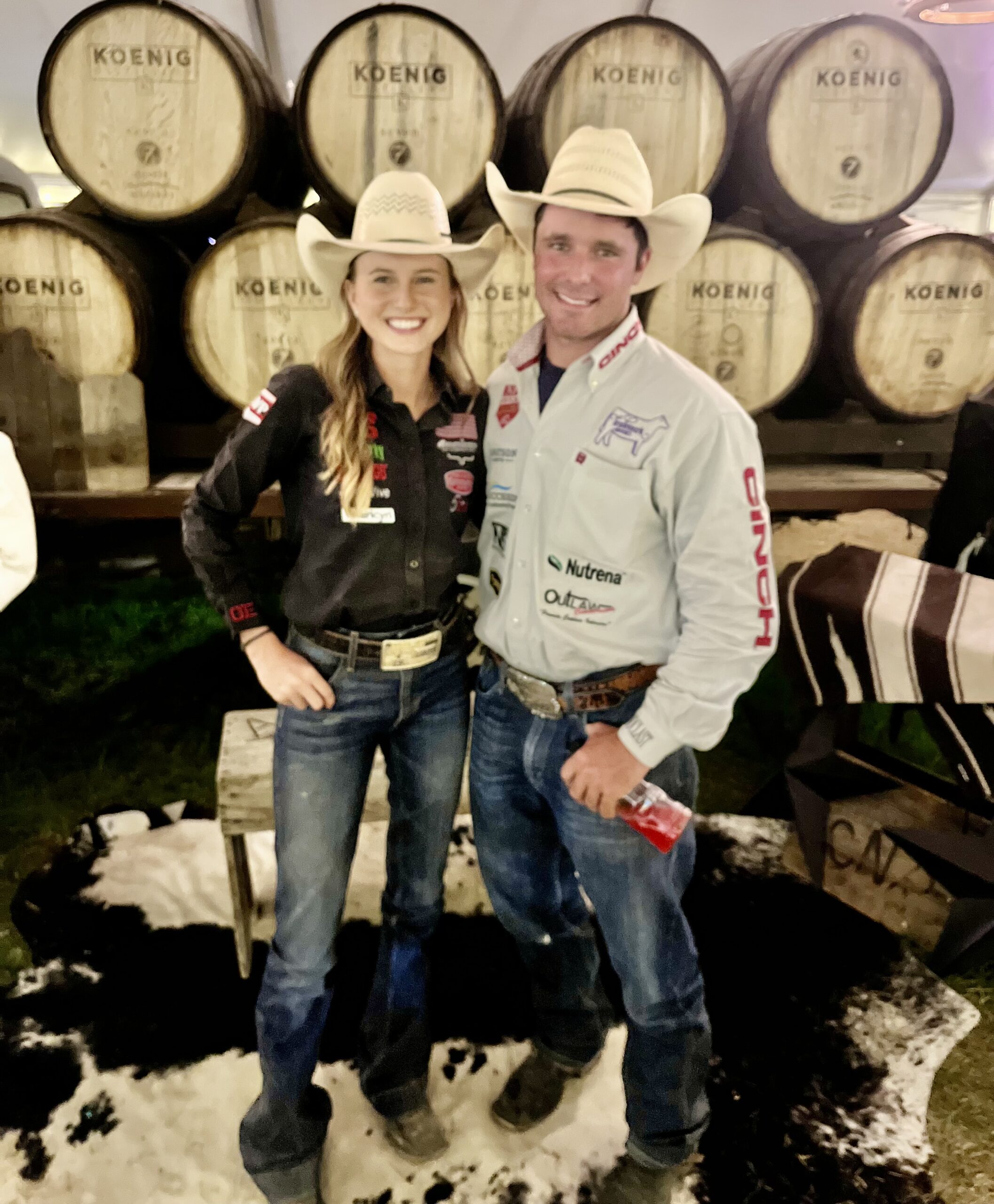 Two smiling rodeo competitors in cowboy hats and sponsor shirts pose arm in arm in front of stacked oak barrels