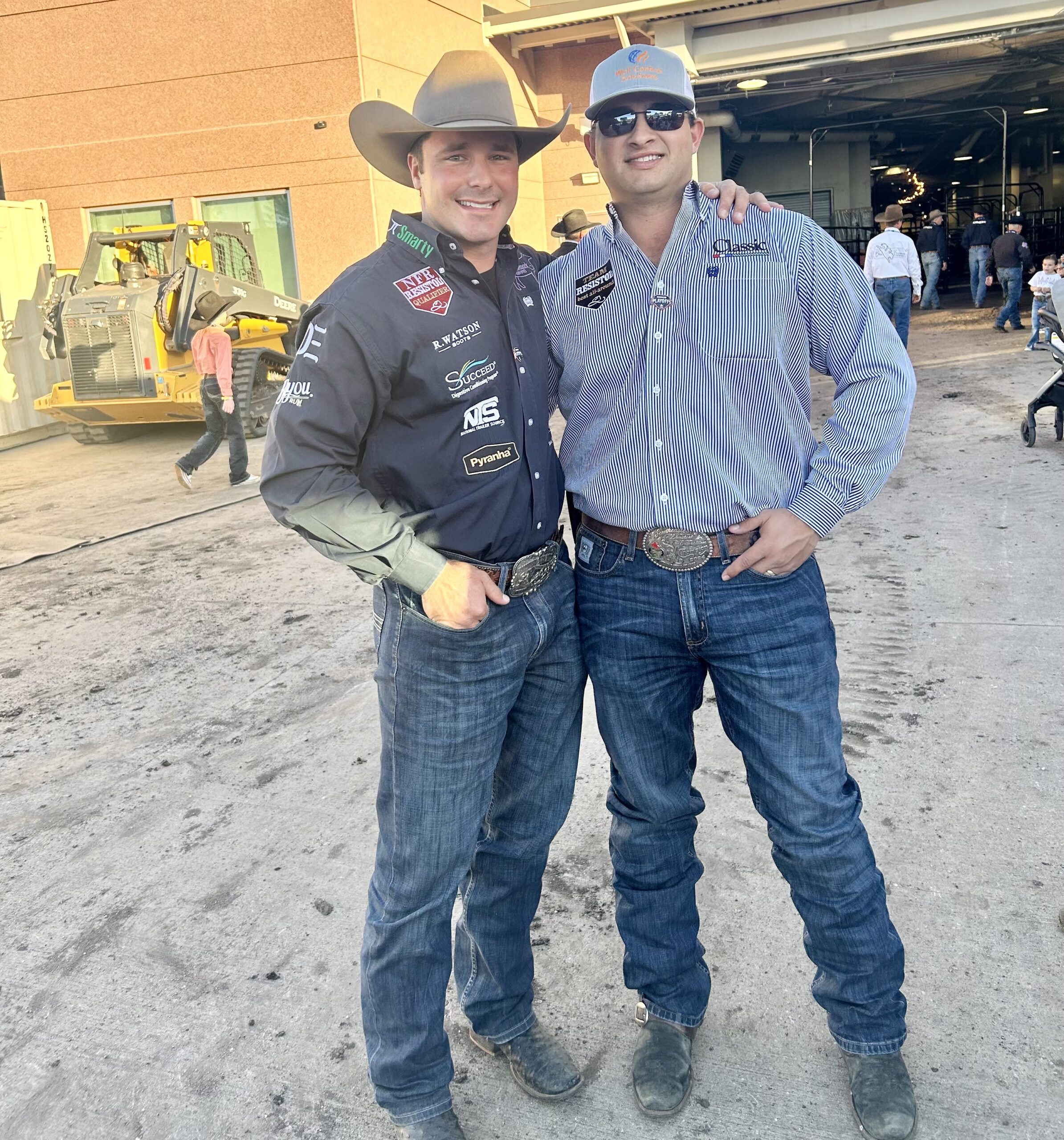 Two smiling cowboys in sponsor-covered western shirts and trophy buckles pose arm-in-arm outside a rodeo arena tunnel
