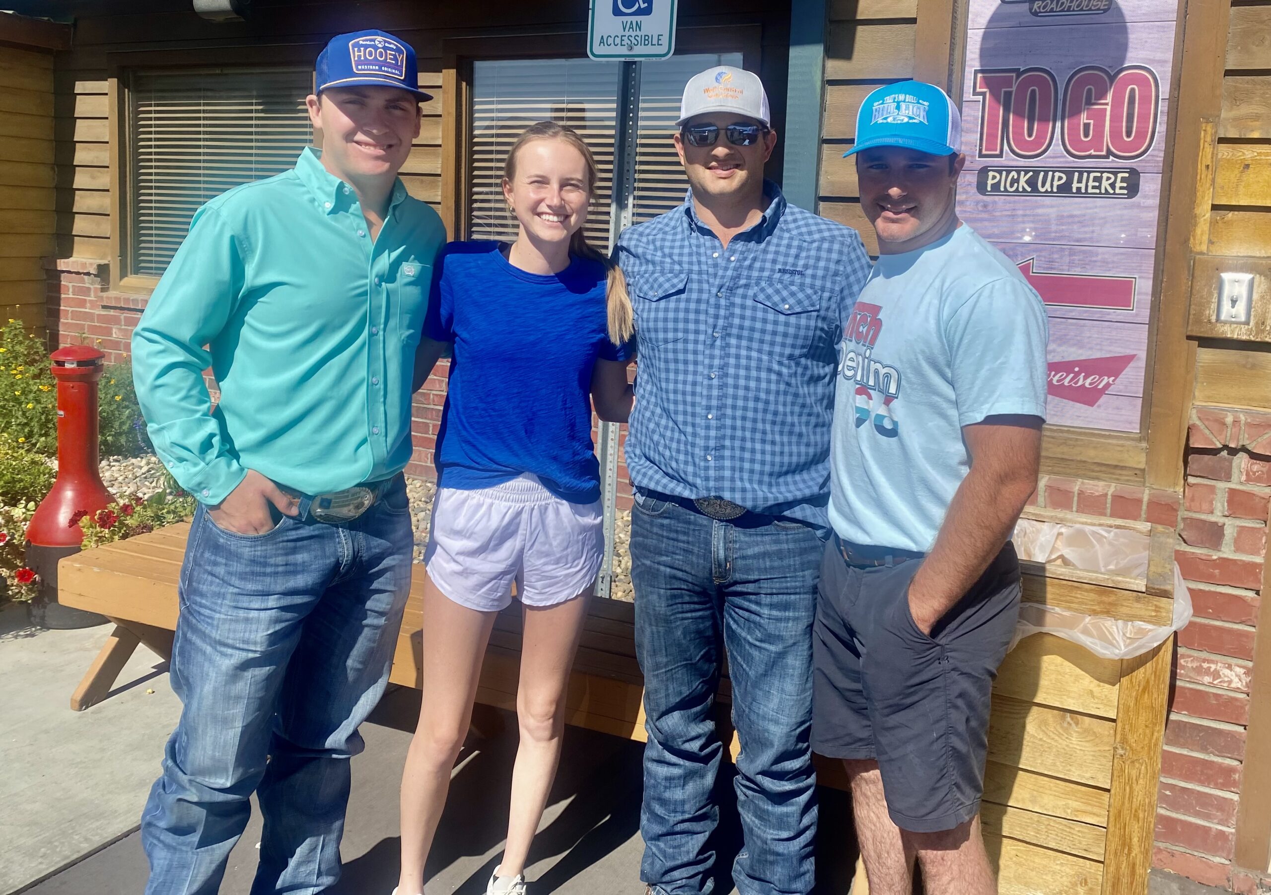 Three men in ball caps and a smiling woman pose arm-in-arm outside a roadhouse restaurant's to-go entrance
