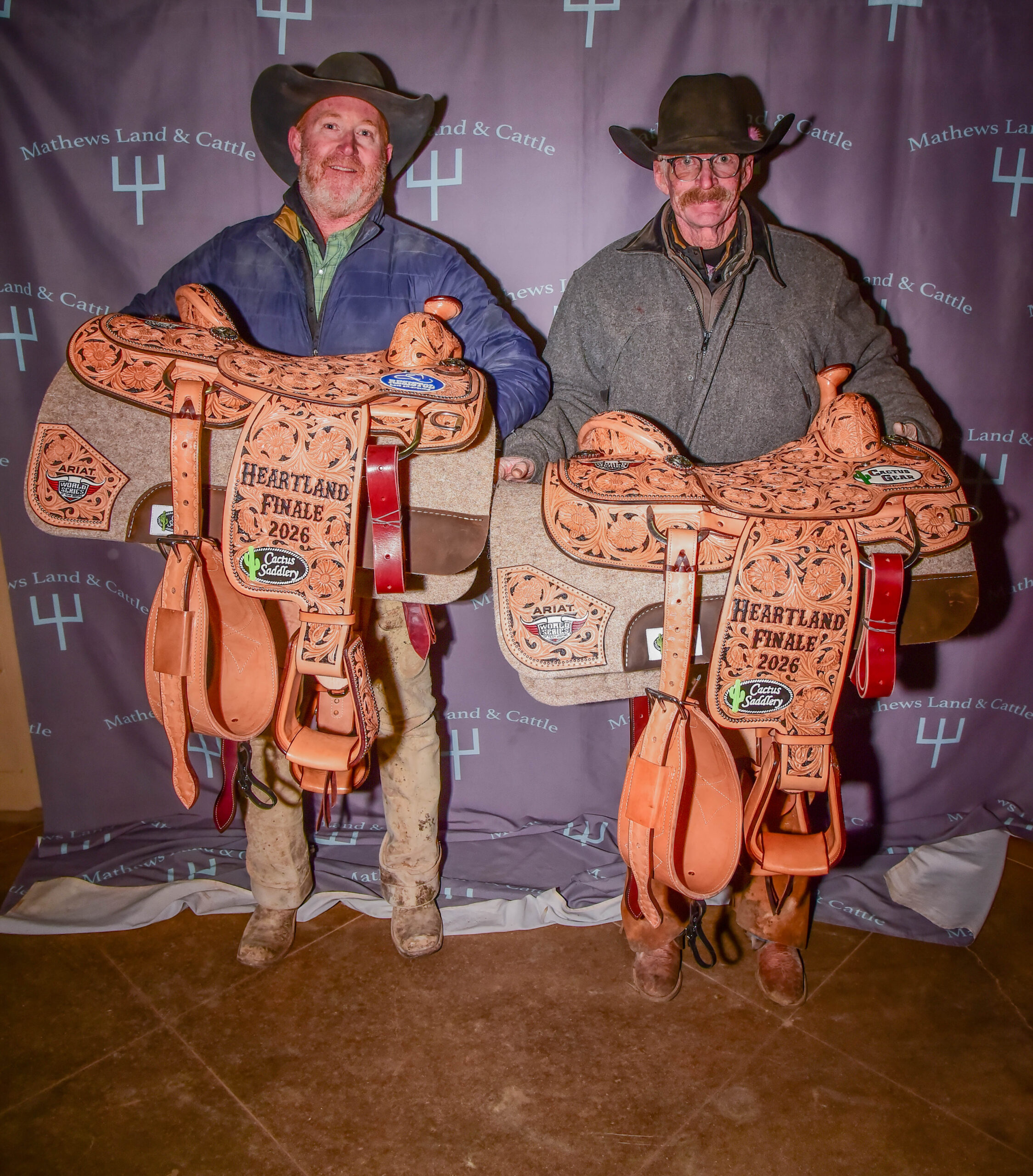 Two cowboys in hats hold tooled Cactus Saddlery "Heartland Finale 2026" champion saddles before a Mathews Land & Cattle backdrop
