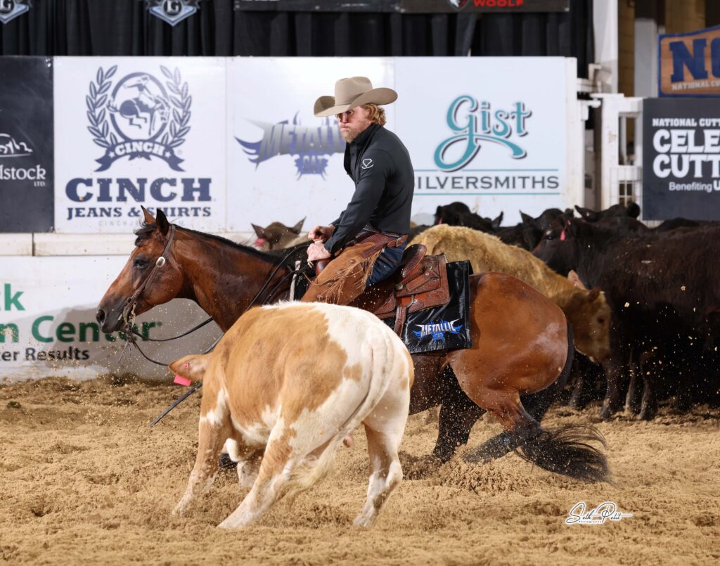 Cutting horse rider in a tan cowboy hat works a tan-and-white cow in the arena as the herd bunches behind