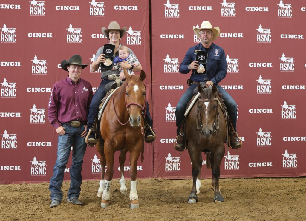 Two mounted riders holding buckle awards, one with a baby in the saddle, beside a standing cowboy at a Cinch RSNC backdrop
