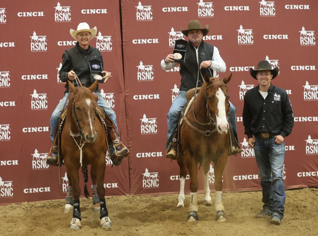 Two mounted cowboys hold up championship buckles beside a standing man in front of a Cinch RSNC backdrop