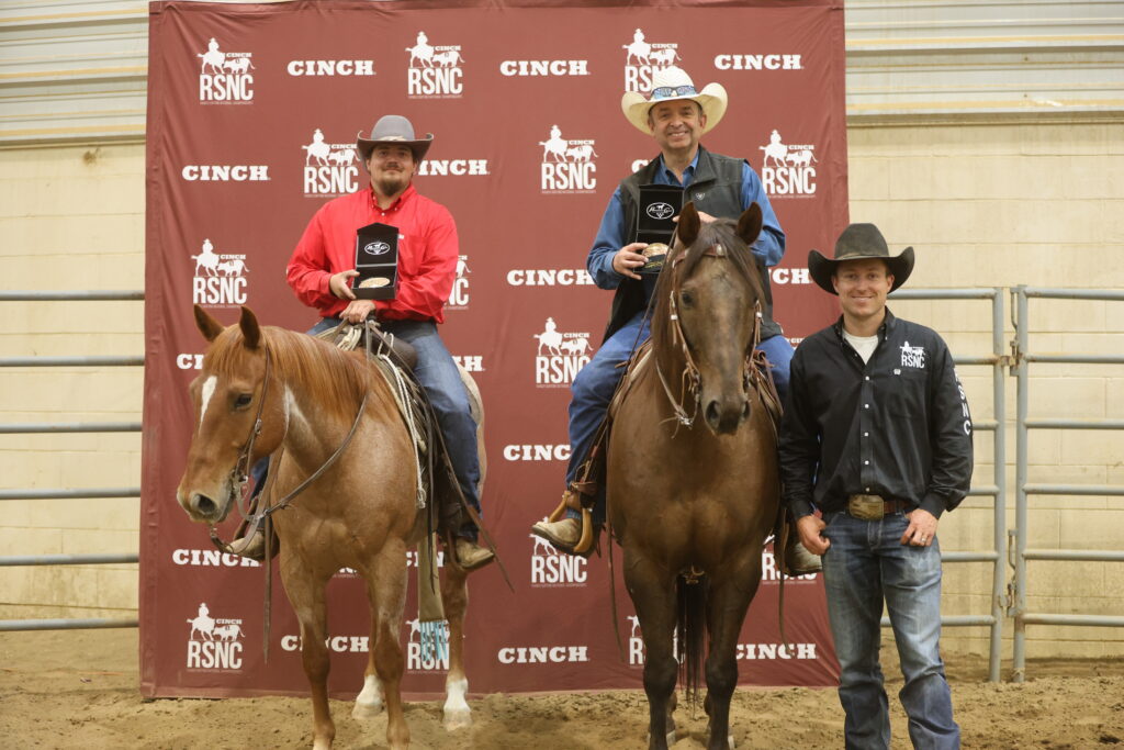 Two mounted riders holding award buckles pose with a standing man in front of a Cinch RSNC backdrop