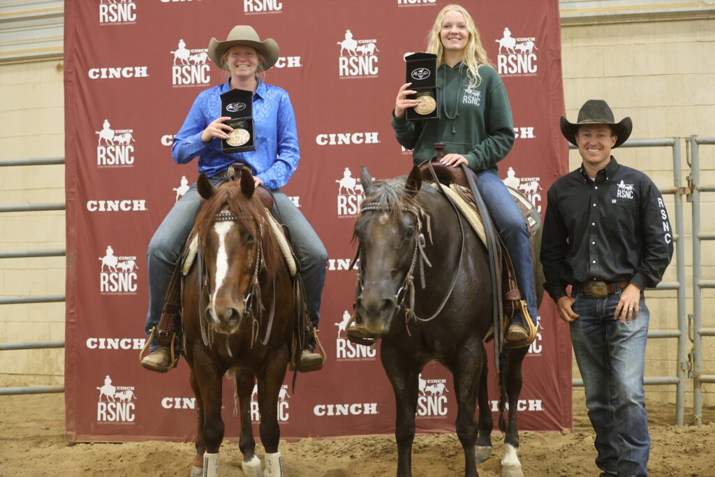 Two mounted riders hold up championship buckles beside a man in a cowboy hat in front of a Cinch RSNC backdrop
