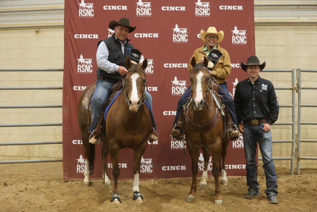 Two mounted riders hold award plaques beside a standing man in front of a Cinch RSNC backdrop in an indoor arena