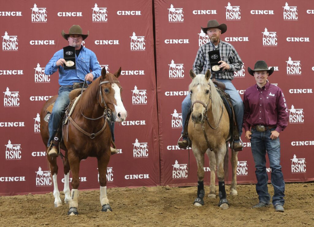 Two mounted cowboys hold up award buckles beside a man standing on the ground in front of a Cinch RSNC backdrop