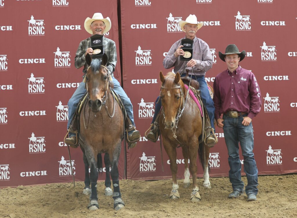 Two mounted cowboys hold up championship belt buckles beside a man standing in front of a Cinch RSNC backdrop