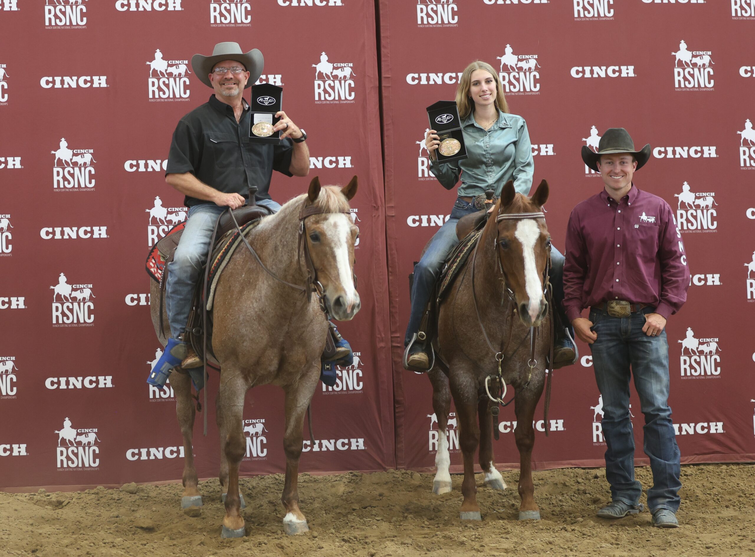 Two mounted ranch sorting winners hold up championship buckles beside a standing official in front of a Cinch RSNC backdrop