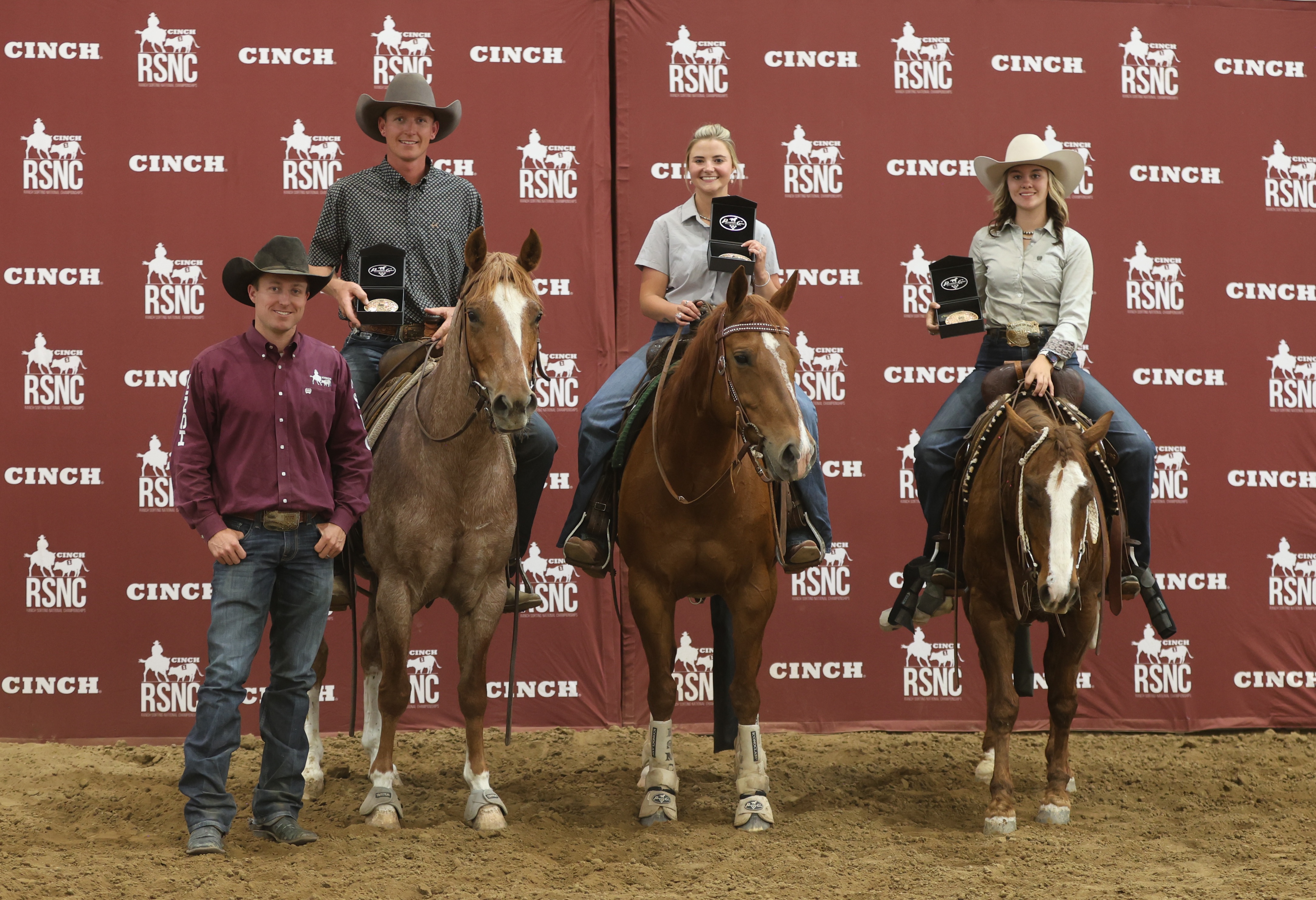 Three mounted riders hold championship buckles in presentation boxes beside a man standing on the ground in front of a Cinch RSNC backdrop