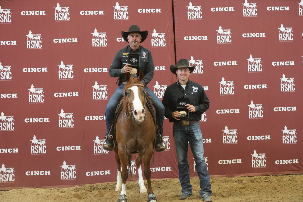 Two men in cowboy hats hold trophy buckles, one mounted on a sorrel horse, in front of a Cinch RSNC backdrop