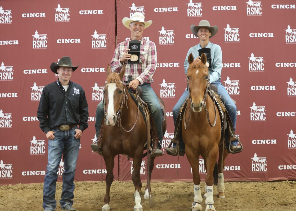 Two mounted riders hold up belt buckle awards beside a standing man in a cowboy hat in front of a Cinch RSNC backdrop