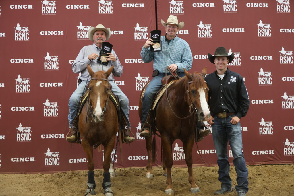 Two mounted riders hold up championship buckles beside a man standing on the ground in front of a Cinch RSNC backdrop