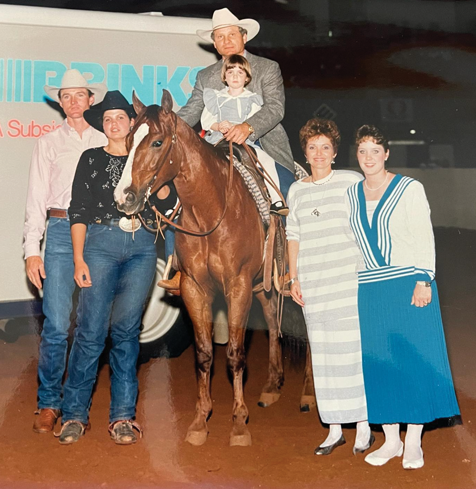 Winner's circle photo of a man in a cowboy hat holding a young girl aboard a sorrel horse, flanked by four people on the arena dirt