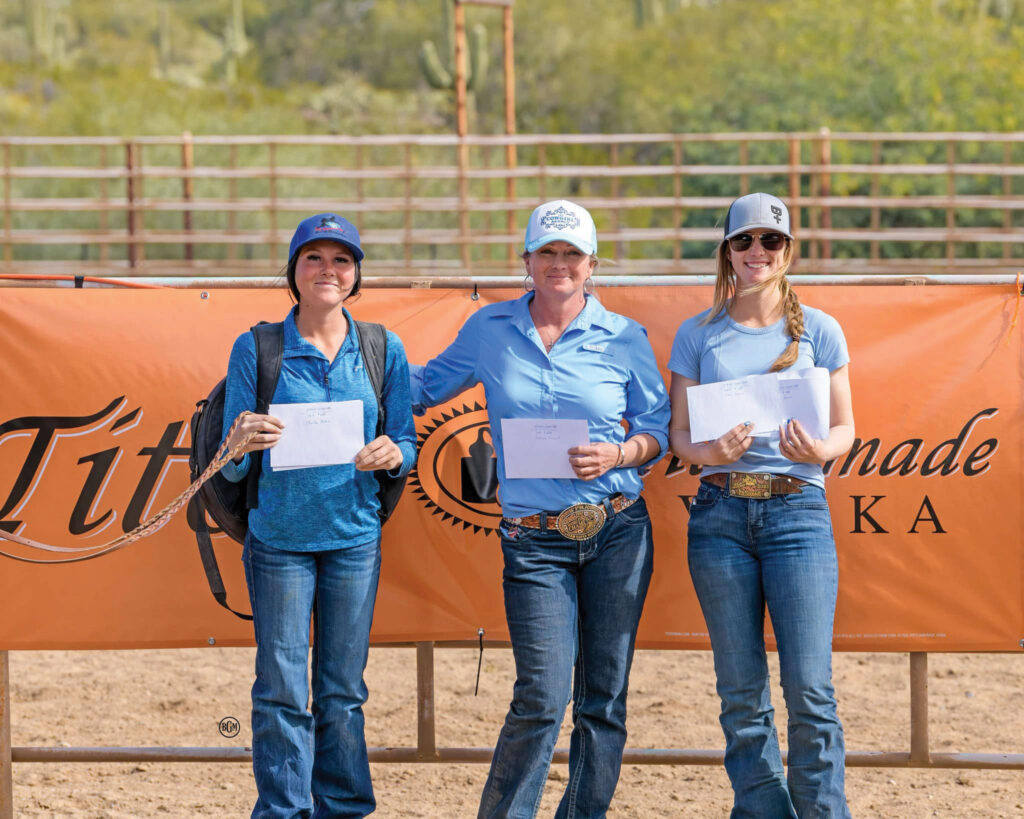 Three women in ball caps hold prize envelopes while standing in an arena in front of an orange sponsor banner