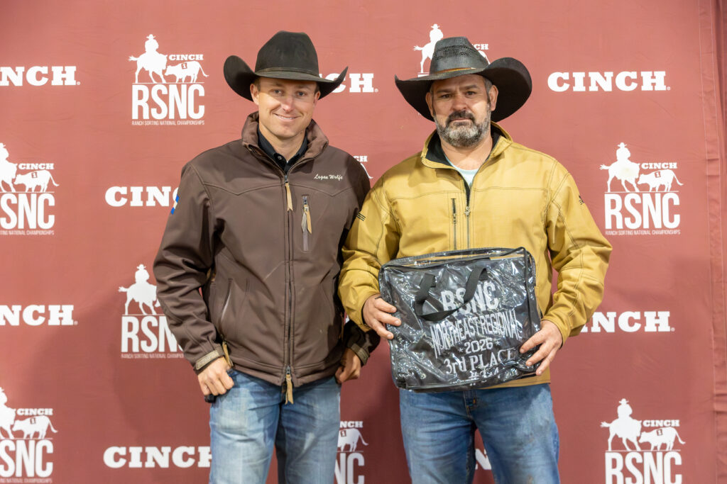 Two cowboys in hats and jackets hold an award bag in front of a Cinch RSNC backdrop after a ranch sorting win