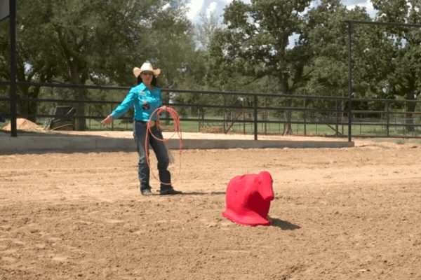 Cowgirl in turquoise shirt and white hat practices roping with a red dummy in a dirt arena