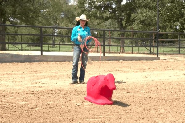 Cowgirl in turquoise shirt and tan hat practices roping with a red dummy in sandy arena with metal fence and trees in background