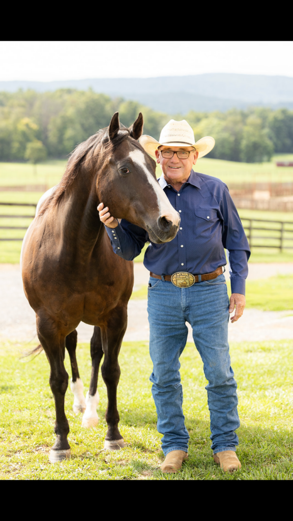 Smiling man in a straw cowboy hat, blue western shirt and NCHA trophy buckle stands beside a dark bay horse in a grassy pasture