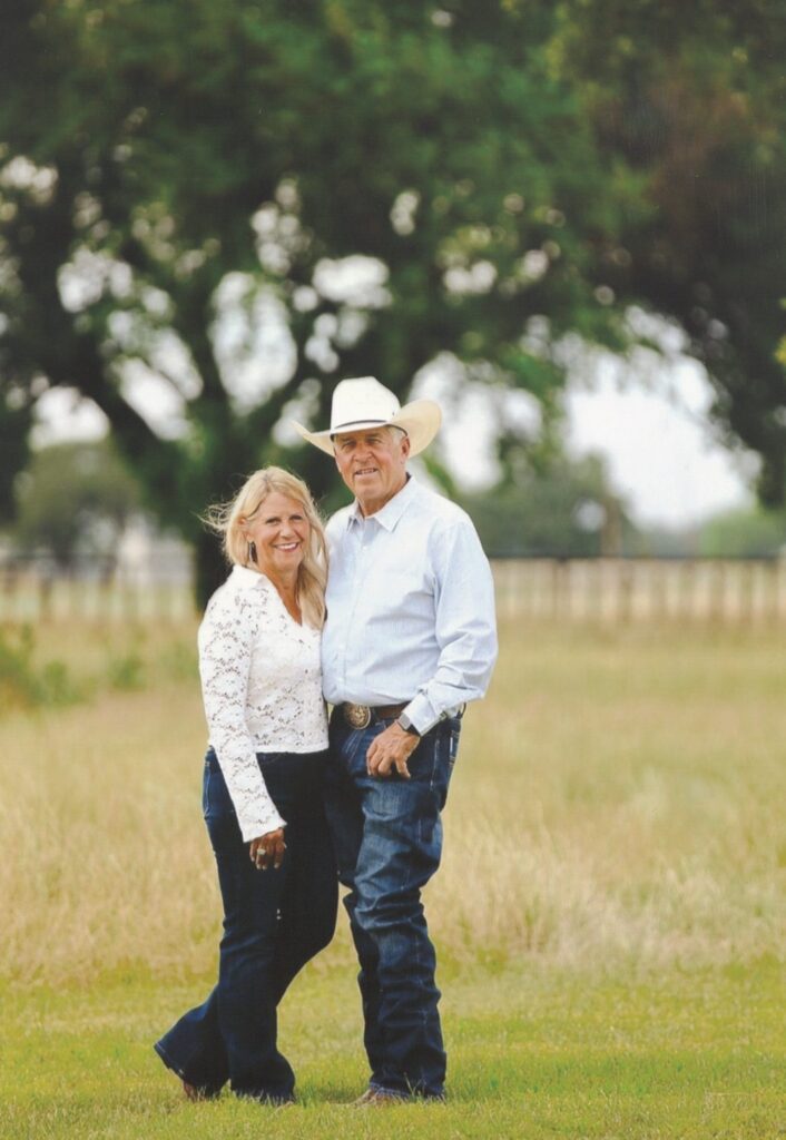 Smiling couple standing arm in arm in a grassy pasture, the man in a white cowboy hat and trophy buckle