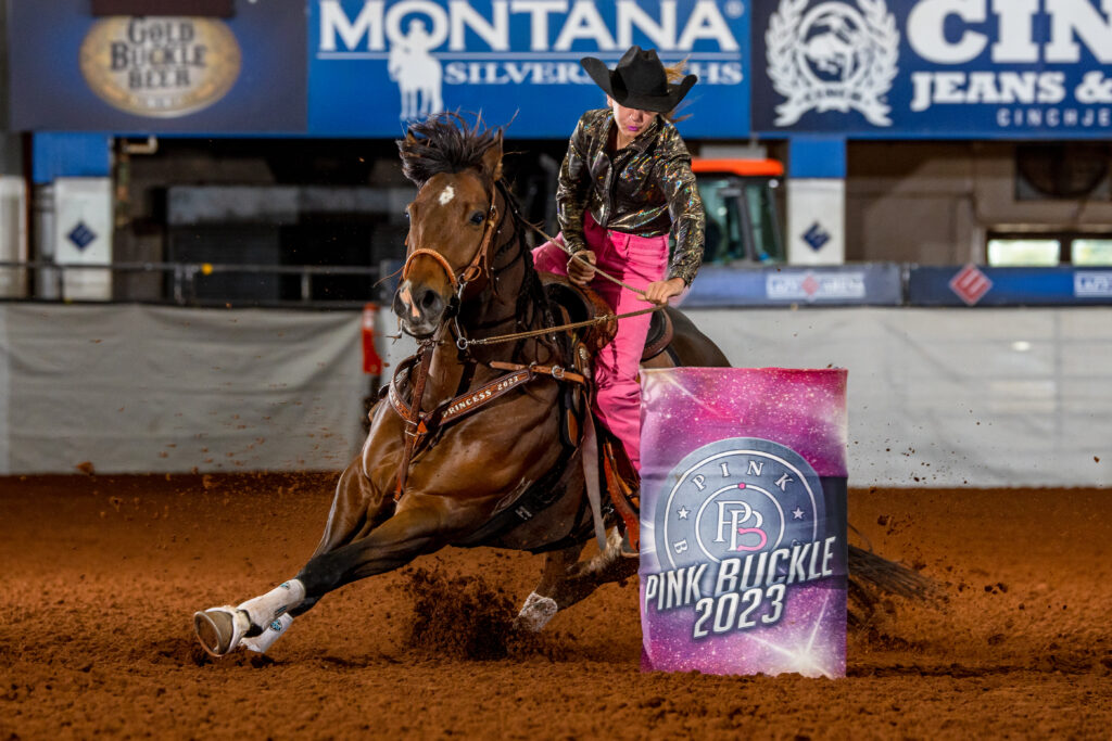 Barrel racer in a black hat and pink pants rounds a Pink Buckle 2023 barrel as her bay horse digs into the dirt