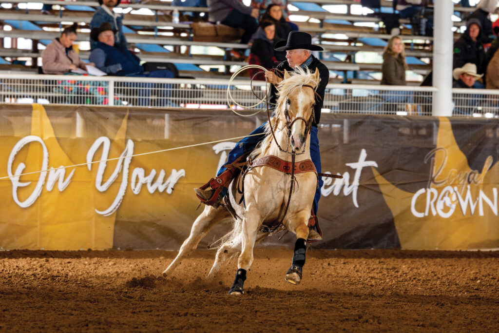 Roper on palomino horse swings lasso during breakaway roping event at indoor rodeo arena with spectators in bleachers