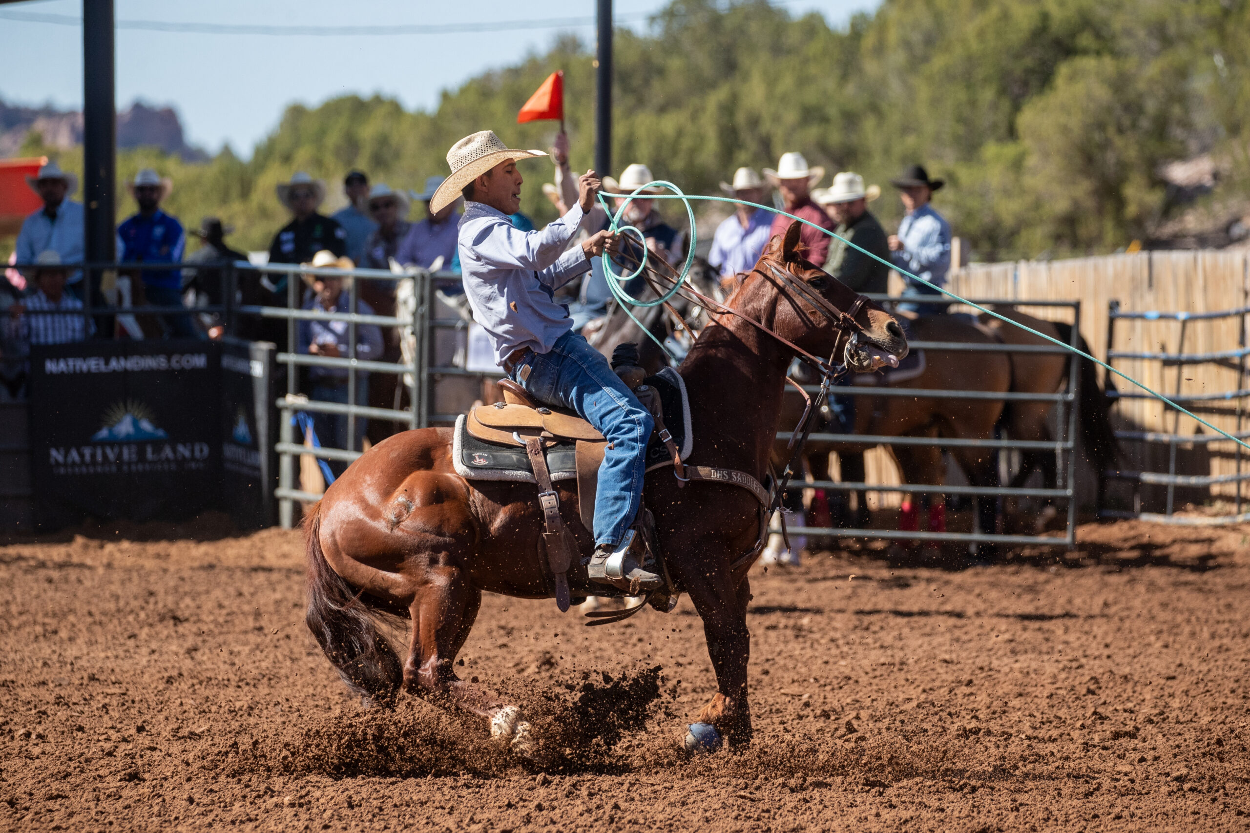 Cowboy on horseback swinging a lasso during a roping event at a rodeo arena with spectators and red flag in background