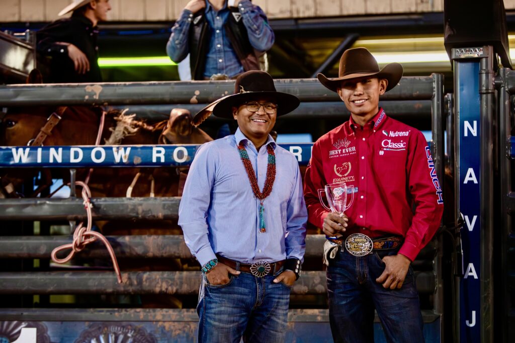 Two rodeo competitors in Western attire stand at a livestock pen, one wearing a red shirt with a belt buckle and holding a trophy