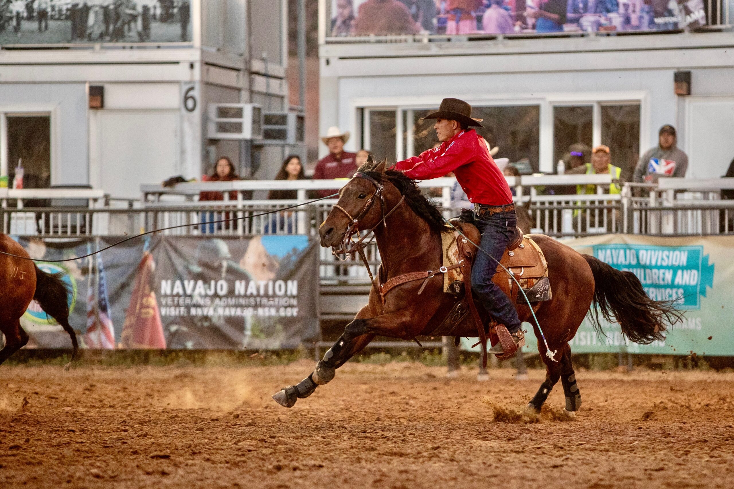 Cowboy in red shirt and brown hat rides brown horse at lope across dirt arena during Navajo Nation rodeo event
