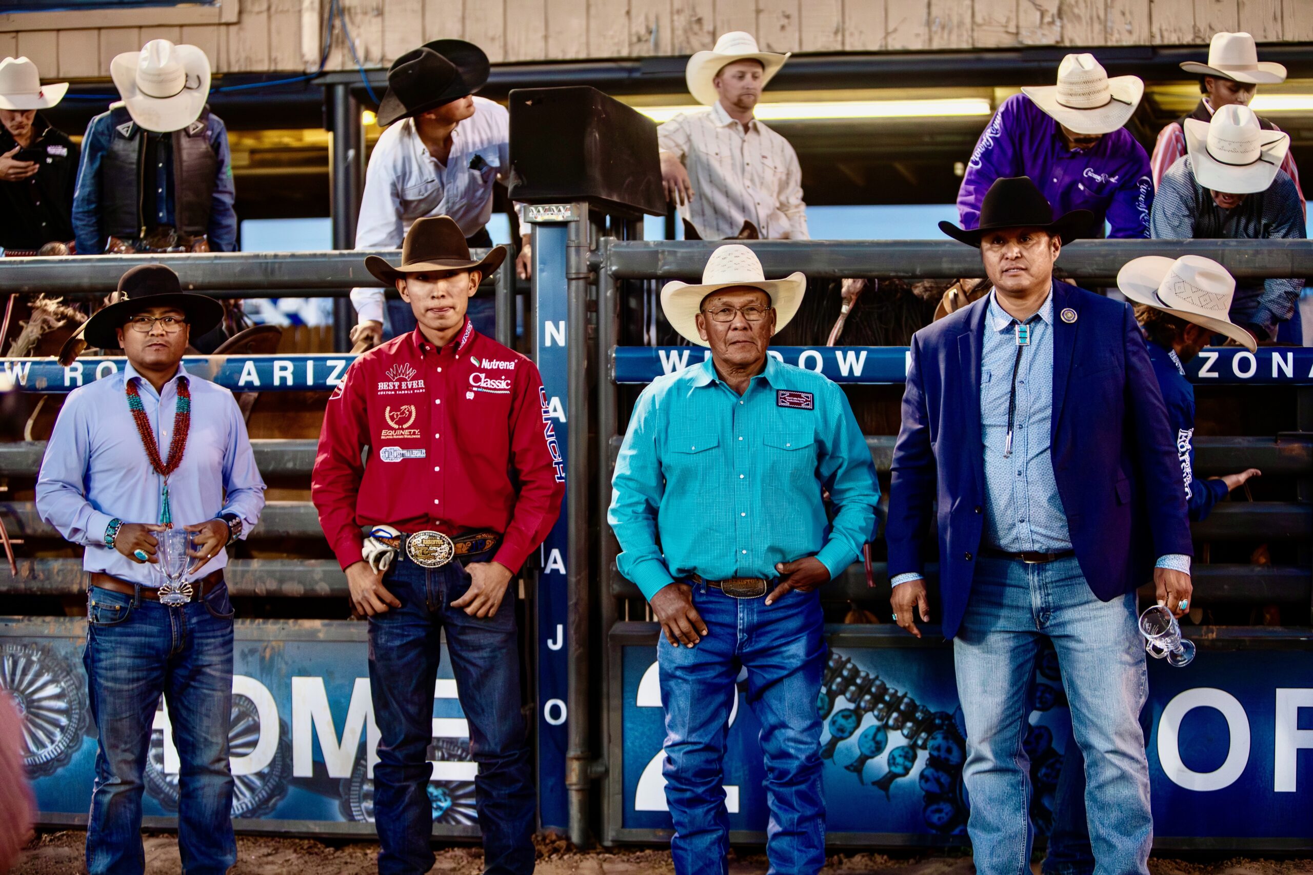 Rodeo officials and dignitaries stand in the arena box at a professional rodeo event, wearing western attire and cowboy hats