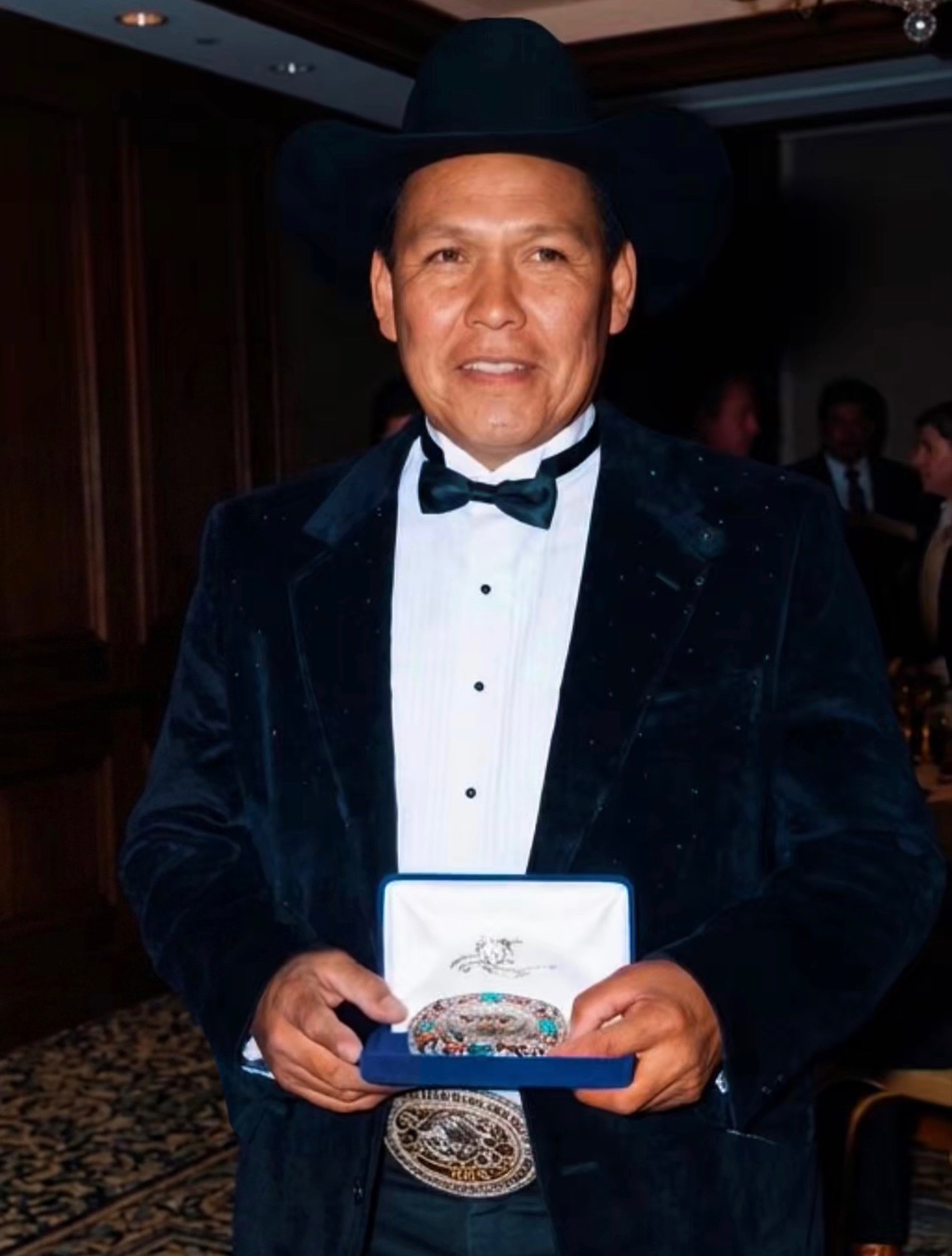 Man in formal Western attire with cowboy hat holding an ornate buckle award at an indoor event