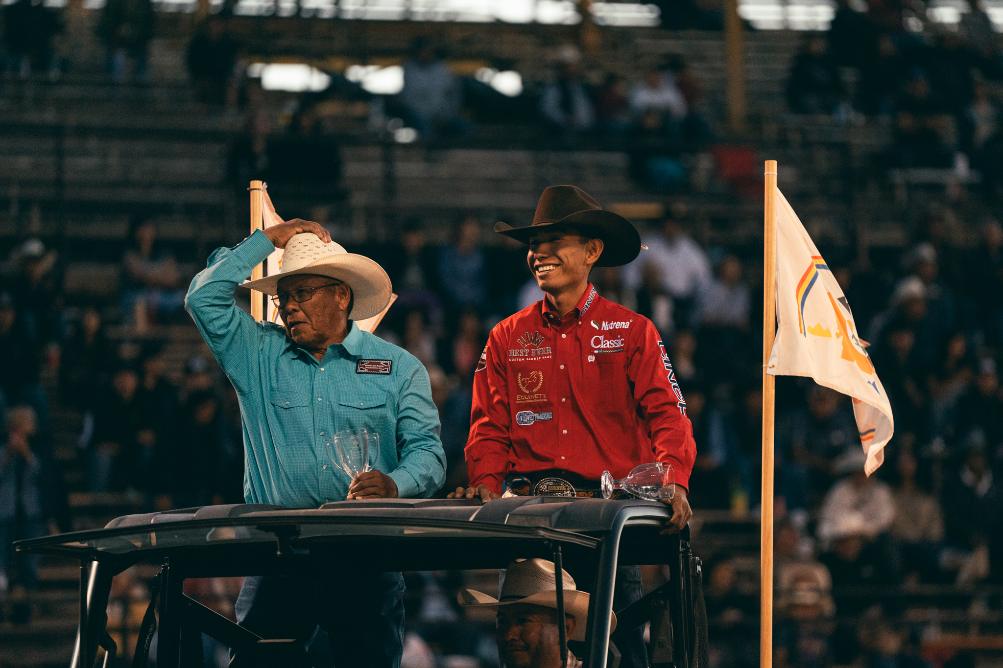 Two cowboys in traditional western attire stand in a vehicle at a rodeo arena during a presentation, with spectators visible in the bleachers behind them