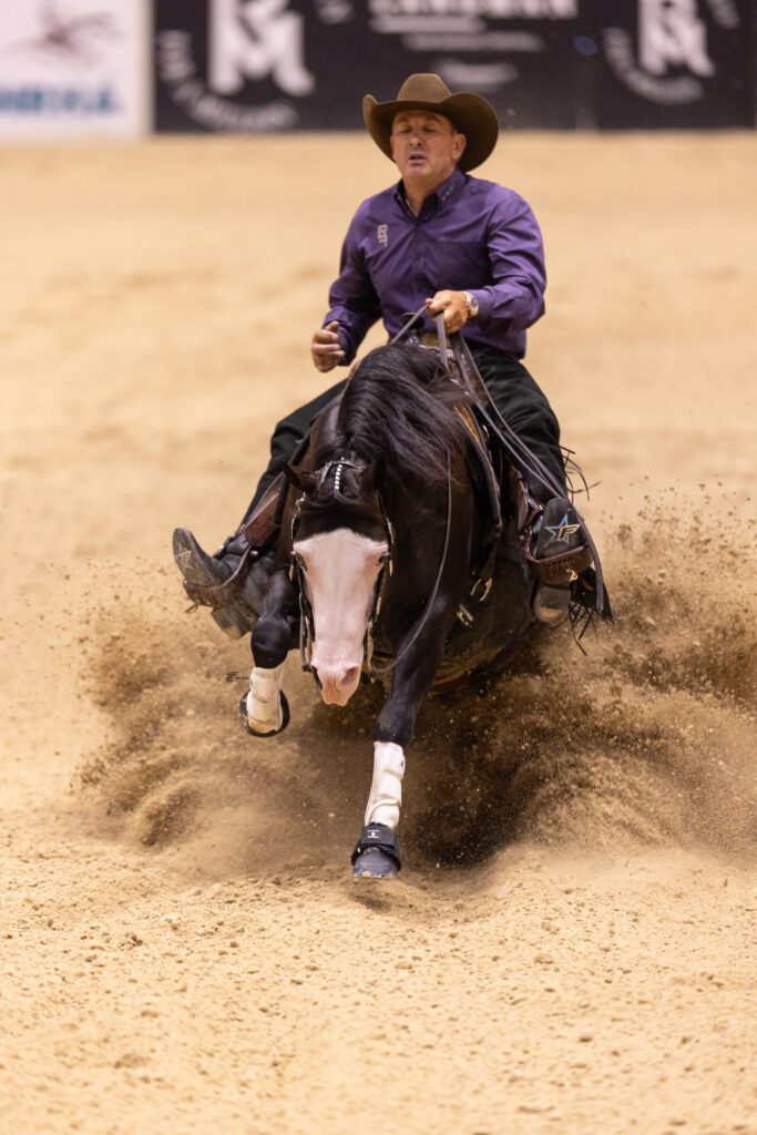 Reiner in a purple shirt and cowboy hat slides his dark horse to a stop, spraying dirt across the arena