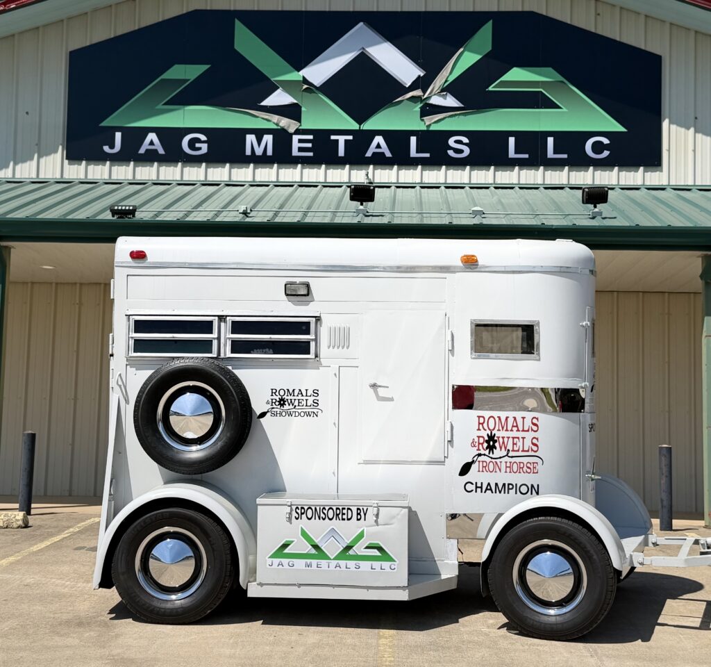 White two-horse trailer with Romals & Rowels Iron Horse Champion decals parked outside the JAG Metals LLC building