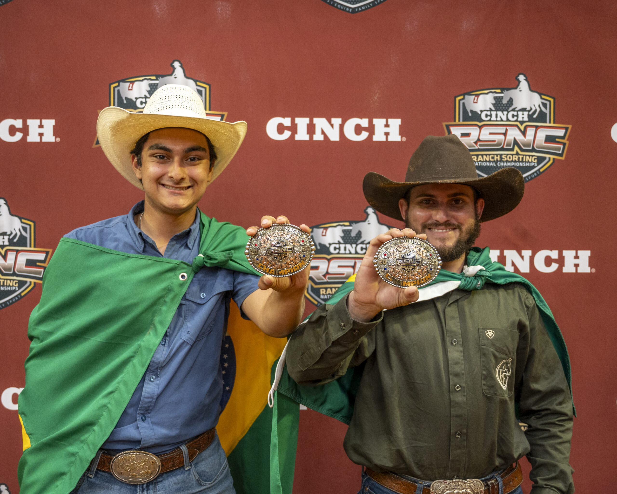 Two cowboys in traditional Western attire display ornate belt buckles at the Cinch RSNC Ranch Sorting National Championships backdrop