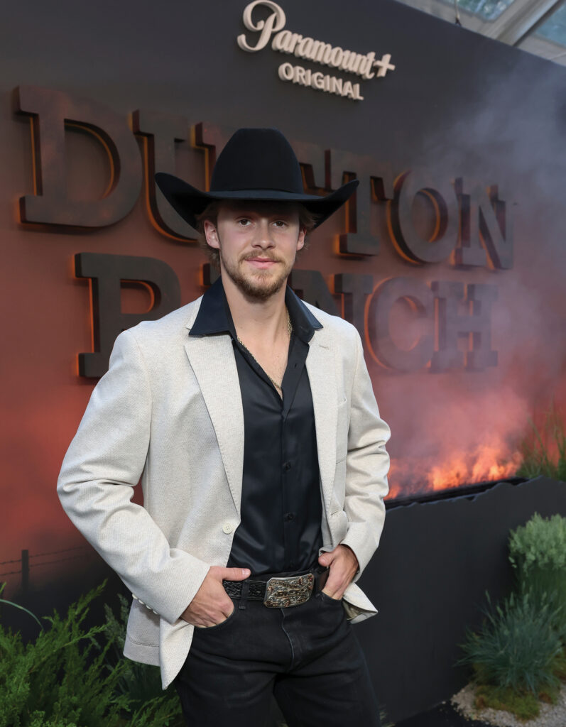 Young man in a black cowboy hat, black shirt, cream blazer and trophy buckle poses on a Western series premiere red carpet