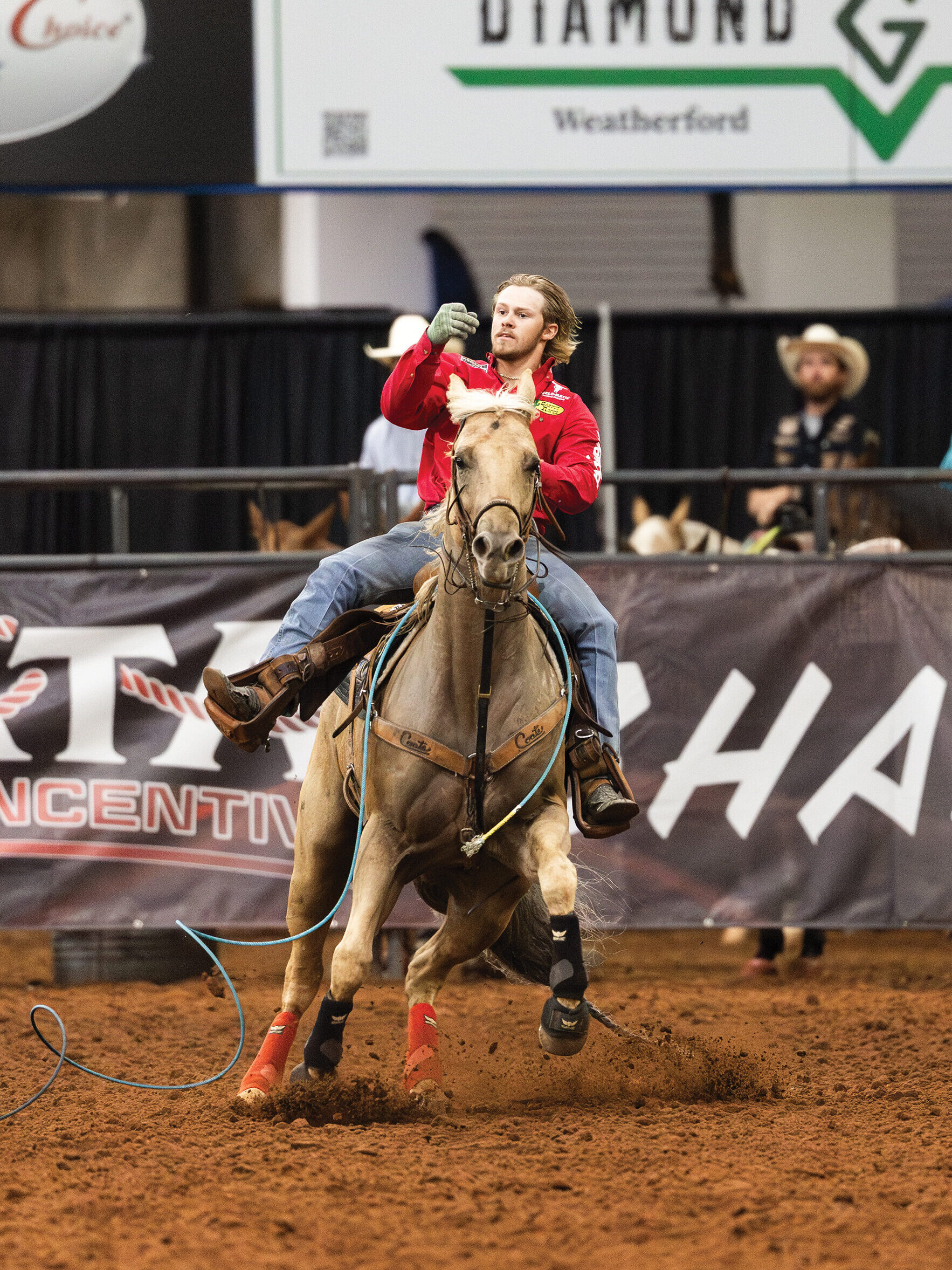 Roper in a red shirt raises his gloved hand after throwing a blue rope from a galloping palomino in an indoor arena