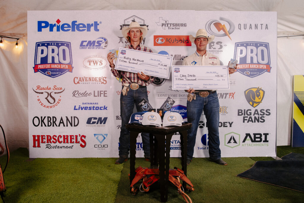 Two rodeo competitors hold large checks at the Priefert Ranch Open event, standing in front of a sponsor backdrop