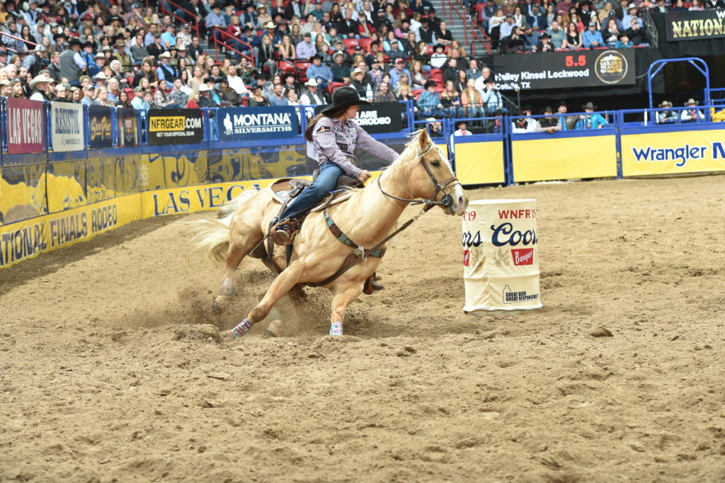 Barrel racer in a black hat turns her palomino hard around a Coors barrel, sand flying, at the National Finals Rodeo