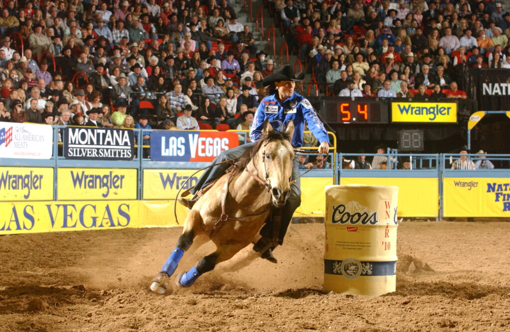 Barrel racer in a blue shirt and black hat rounds a Coors barrel on a buckskin horse as the clock reads 5.4 at an indoor rodeo